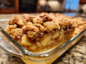 A close-up shot of a slice of warm apple pie dump cake in a glass baking dish, showing tender apple filling and a crumbly topping.