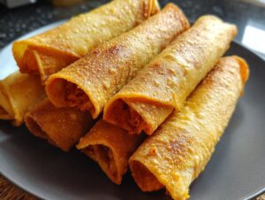 A close-up shot of golden-brown baked chicken taquitos stacked on a dark plate, showing their crispy texture.