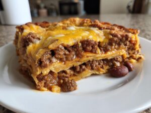 A close-up of a hearty slice of Beef Taco Casserole on a white plate, showing layers of seasoned ground beef, tortillas, and melted cheese.