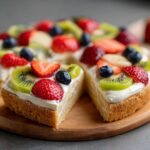 Close-up of slices from a fruit pizza sugar cookie, topped with cream cheese frosting and fresh berries and kiwi.