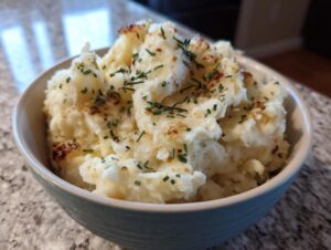 A close-up of a bowl filled with creamy garlic parmesan mashed potatoes, topped with chives and browned bits.