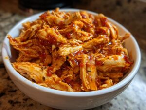A white bowl filled with shredded slow cooker sweet chili chicken, coated in a glossy, reddish-orange sauce.