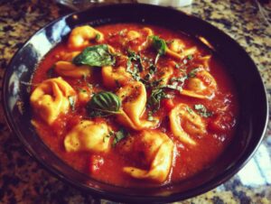 A close-up of a black bowl filled with delicious tomato basil tortellini soup, garnished with fresh basil leaves.
