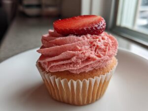 A close-up of a single, perfectly frosted strawberry cupcake topped with a fresh strawberry slice.