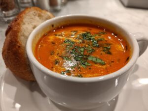 A close-up of a creamy, vibrant tomato bisque topped with fresh basil and cream swirl, served with a slice of bread.