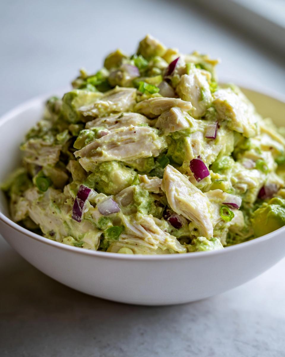 Close-up of creamy avocado chicken salad featuring shredded chicken, chunks of avocado, and diced red onion in a white bowl.