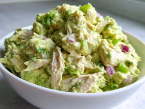 A close-up of creamy avocado chicken salad mixed with red onion and celery served in a white bowl.