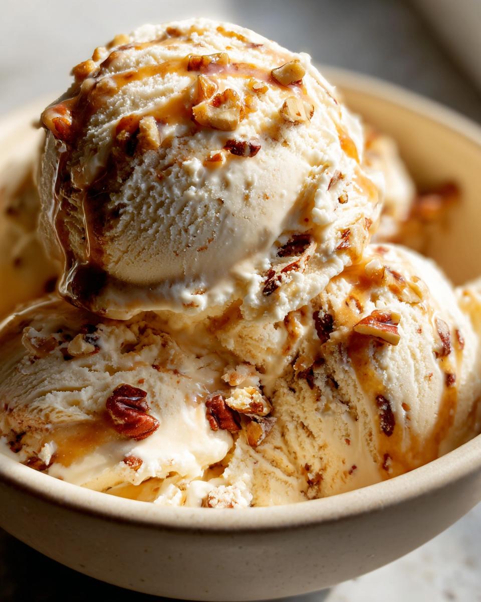 Close-up of creamy butter pecan ice cream scoops in a bowl, drizzled with caramel and topped with chopped pecans.