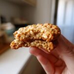 A hand holding a freshly baked oatmeal cookie broken in half, showing its chewy texture.