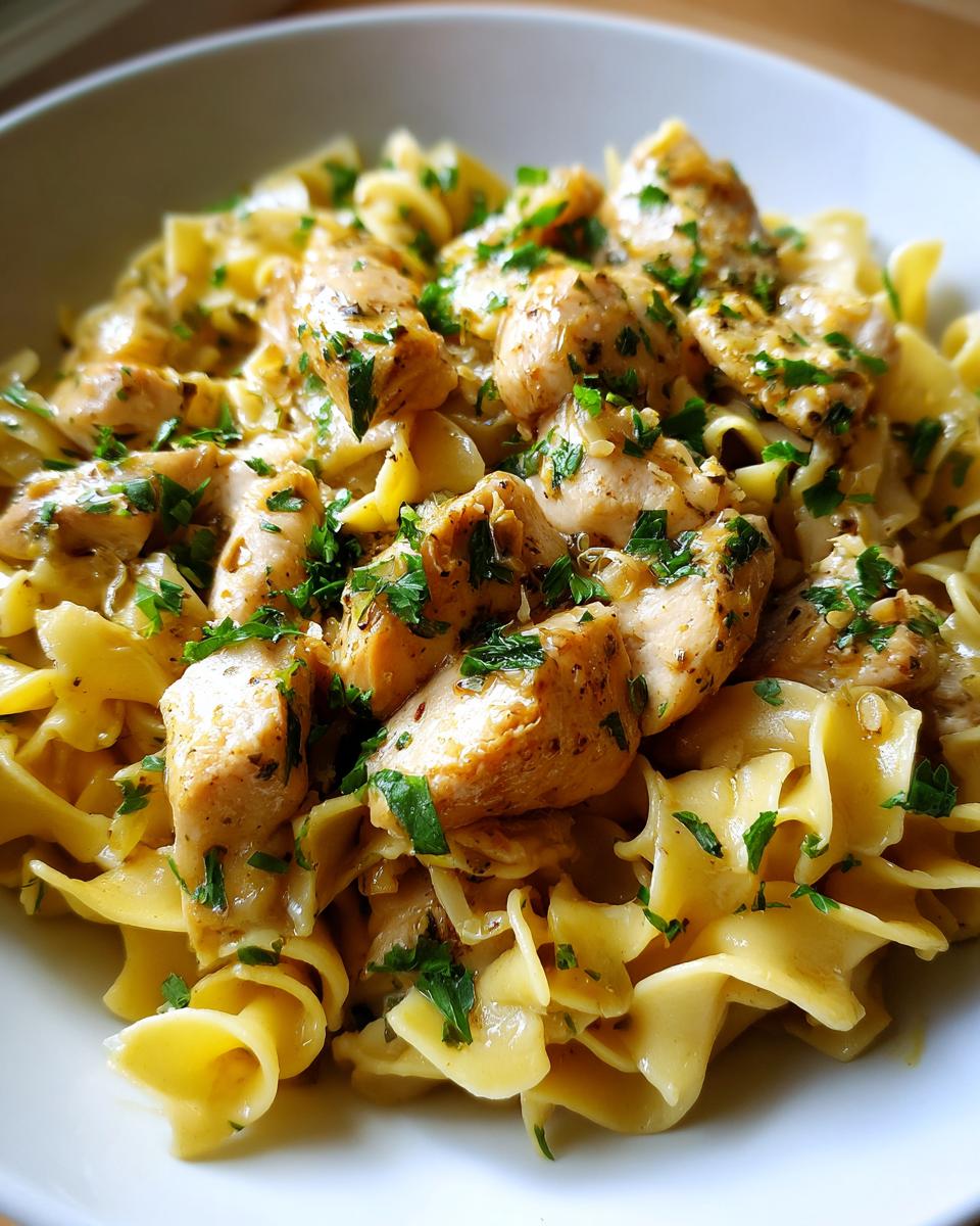 A close-up of tender chicken pieces served over wide egg noodles, garnished with fresh parsley. This is chicken with buttered noodles.