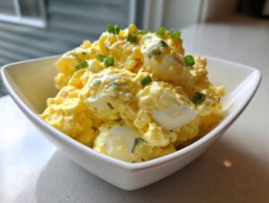 A close-up of creamy, yellow egg salad garnished with chopped green onions in a modern white serving bowl.
