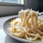 Close-up of fettuccine noodles coated in thick homemade alfredo sauce being lifted by a fork, topped with grated Parmesan cheese.