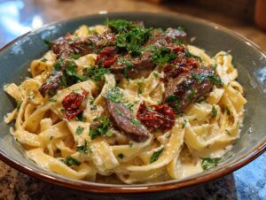 A bowl of creamy steak pasta with fettuccine, sliced steak, sun-dried tomatoes, and parsley.