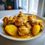 A close-up of rich, yellow Jamaican curry chicken pieces served with large chunks of potato in a white bowl.