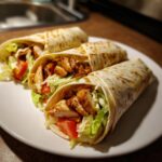 Three delicious chicken wraps filled with seasoned chicken, lettuce, tomato, and sauce on a white plate.