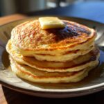 A stack of three fluffy old fashioned pancakes, topped with a pat of melting butter and drizzled with syrup.