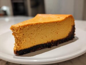 A close-up slice of rich, orange pumpkin cheesecake on a white plate showing a dark chocolate crust.