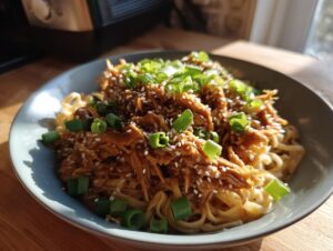 A bowl of slow cooker honey garlic chicken and noodles topped with sesame seeds and green onions.