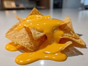 A pile of crispy tortilla chips generously covered in bright, smooth nacho cheese sauce dripping onto a white counter.