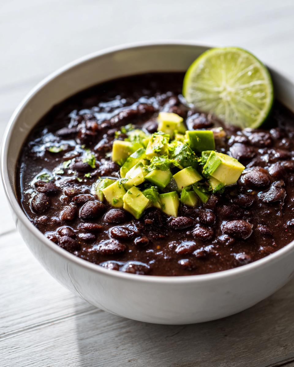 Close-up of a white bowl filled with rich black bean soup, topped with diced avocado and a lime wedge.
