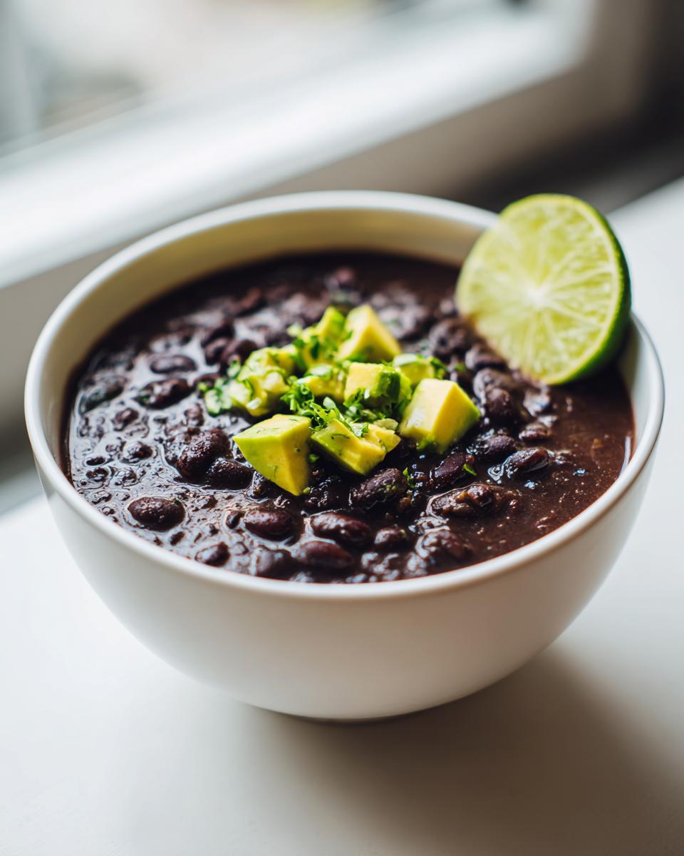 A close-up of a bowl of rich black bean soup garnished with diced avocado and a lime wedge.