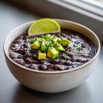 A close-up of a bowl of rich, dark black bean soup topped with diced avocado, cilantro, and a lime wedge.