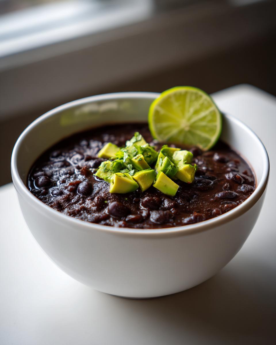 A white bowl filled with rich, dark black bean soup, topped with diced avocado and a lime wedge.