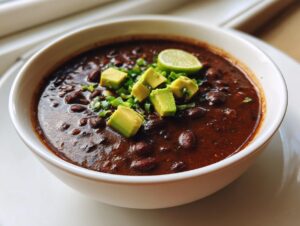 A close-up of a hearty bowl of rich black bean soup garnished with diced avocado, green onions, and a lime wedge.