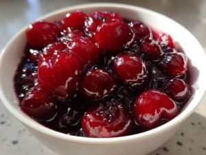 A close-up, bright photo of glossy, whole cherries suspended in thick, dark syrup, representing homemade cherry pie filling.
