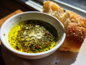 Close-up of a white bowl filled with vibrant olive oil bread dip topped with herbs, spices, and Parmesan, next to crusty bread.