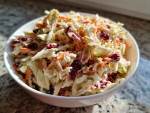 A close-up of a white bowl filled with creamy apple cranberry coleslaw, featuring shredded cabbage, carrots, and dried cranberries.