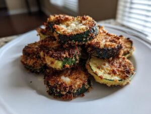 A pile of crispy, golden-brown baked parmesan zucchini slices stacked on a white plate.