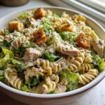 A close-up of a creamy chicken caesar pasta salad featuring rotini pasta, romaine lettuce, chicken pieces, and croutons.