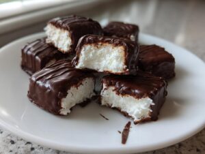 Close-up of several chocolate-covered squares of white, fluffy cool whip candy on a white plate.