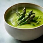 Close-up of a steaming bowl of vibrant green, creamy asparagus soup garnished with whole roasted asparagus spears.