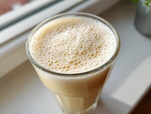 Overhead view of a creamy banana smoothie with a thick layer of foam in a tall glass, sitting on a windowsill.
