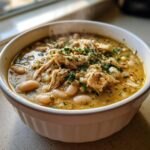 Close-up of a steaming white bowl filled with creamy chicken chili, featuring shredded chicken, white beans, and fresh parsley.