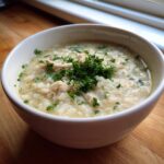 A white bowl filled with creamy rice soup featuring shredded chicken and topped with fresh parsley.
