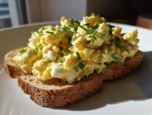 A generous serving of creamy egg salad recipe piled high on two slices of toasted brown bread, topped with fresh chives.