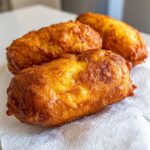 Three freshly fried, golden brown corn dogs resting on a white paper towel to drain.