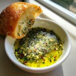 A piece of crusty bread being dipped into a small white bowl of flavorful olive oil bread dip with herbs and cheese.