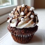 Close-up of a rich chocolate cupcake topped with white frosting, chocolate drizzle, mini marshmallows, and cocoa powder, resembling hot chocolate cupcakes.