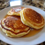 Three golden brown, fluffy discard pancakes stacked slightly on a white plate, covered in syrup.