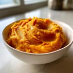 A close-up of creamy, vibrant orange mashed sweet potatoes served in a simple white bowl.