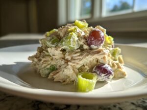 A mound of creamy greek yogurt chicken salad mixed with celery pieces and red grapes, served on a white plate.