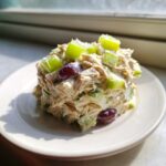 A scoop of creamy greek yogurt chicken salad mixed with green celery chunks and red grapes on a white plate.