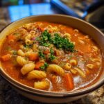 Close-up of a steaming bowl of rich, tomato-based Pasta e Fagioli soup with elbow macaroni, beans, carrots, and parsley.