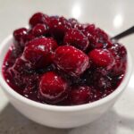 A close-up of glistening, deep red cherry pie filling with whole cherries in a small white bowl.