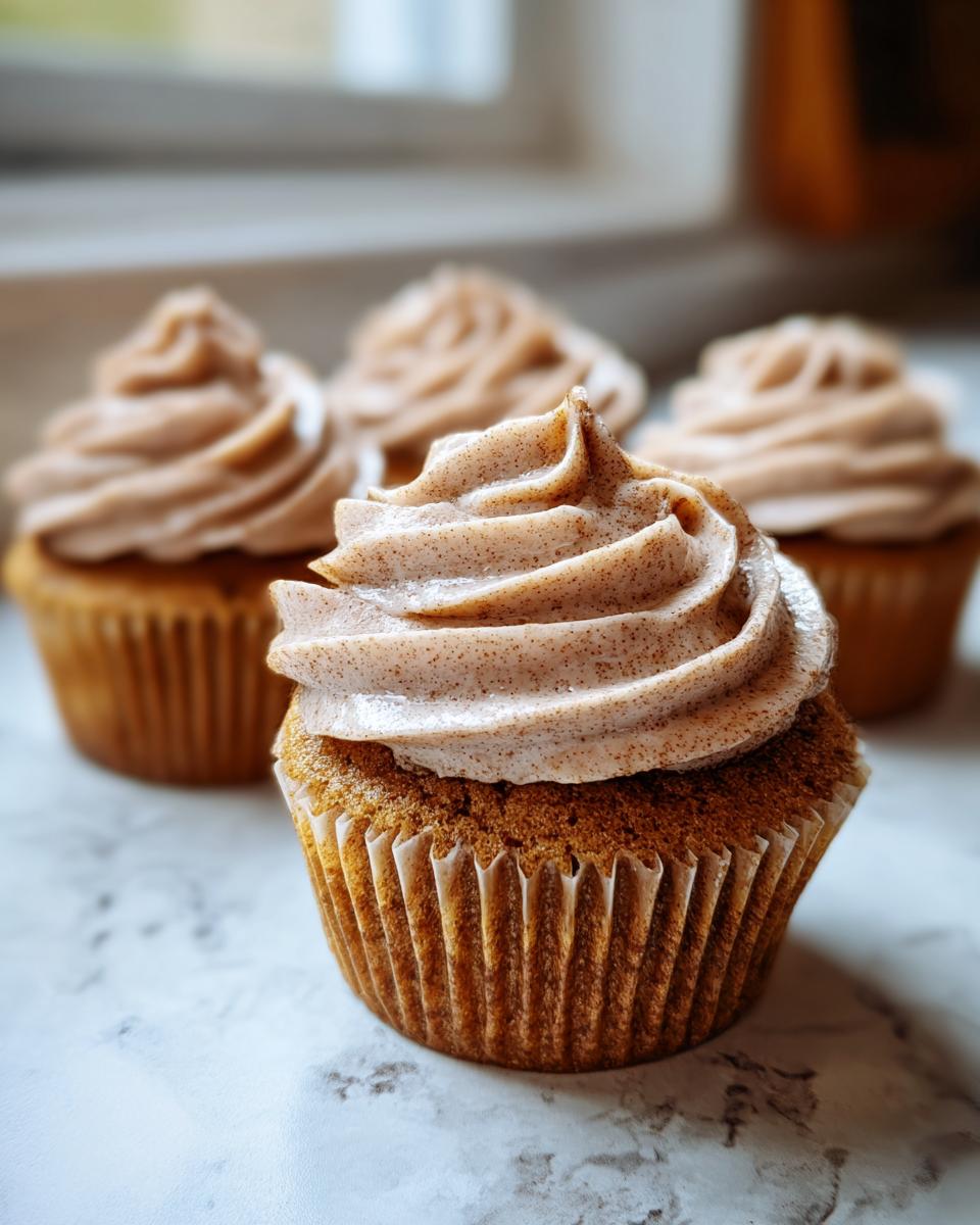A close-up of one moist banana cupcake topped with swirled, speckled cinnamon frosting, with others blurred in the background.