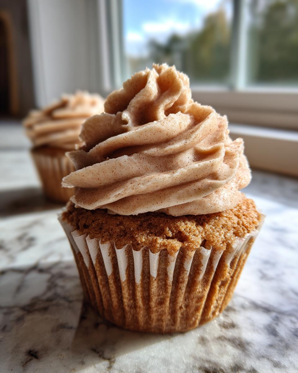 Close-up of a moist banana cupcakes topped with swirled, cinnamon-flecked frosting, sitting on a marble counter.